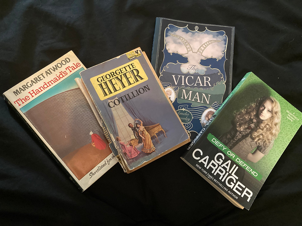 Four books lying, though you can't easily tell from the image, on a bed. The books are The Handmaid's Tale in its original paperback, showing a picture of a high wall and a woman in a shapeless red dress and white, concealing cap; A very battered copy of Georgette Heyer's Cotillion, showing a brunette woman in a yellow dress, dancing with a dark-haired, dark-coated man, while a blonde woman in pink plays the piano, against a lilac background; my own book, The Vicar Man, with part of the cover image covered by the other books, but showing parts of portraits of a blond man and a dark haired woman under the title, and a horned head outlined against the sky above it; and Gail Carriger's Defy Or Defend, showing a blonde woman in dark clothing staring calmly out at the viewer, against a background of green wallpaper. Ostensibly these are here to illustrate the different ways in which we view different books, but mostly I just get a guilty thrill out of seeing my book included. The duvet on which the books recline is black and not perfectly smoothed out.