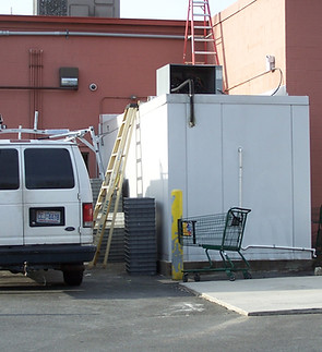 A parked white Ford van is next to a large white utility box with a shopping cart in front of it. A metal ladder extends from the ground to the roof of the box, and a second ladder rests against the side of a red-brick building.