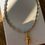 Thumbnail: Silver bead-chain necklace with gold conch-shell pendant laid flat on a light surface and wooden table.
