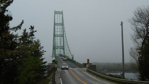 A tall, green bridge rises out of the fog. There is a two lane paved road with a car coming forward