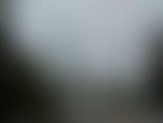 A tall, green bridge rises out of the fog. There is a two lane paved road with a car coming forward