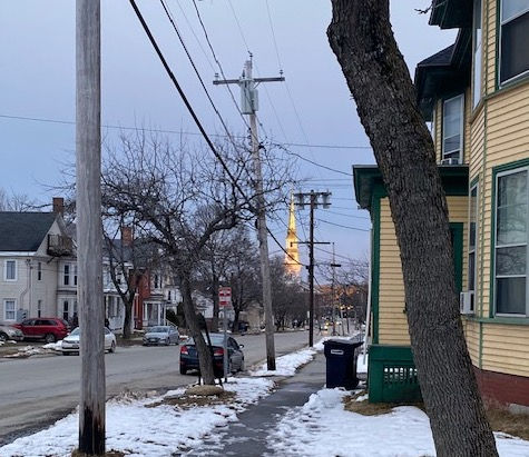 Snowy street scene with bare trees, utility poles, and a yellow house. A distant church spire reflects sunlight. Overcast sky.
