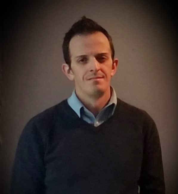 An image of a white man in a brown sweater with a blue shirt on a brown background