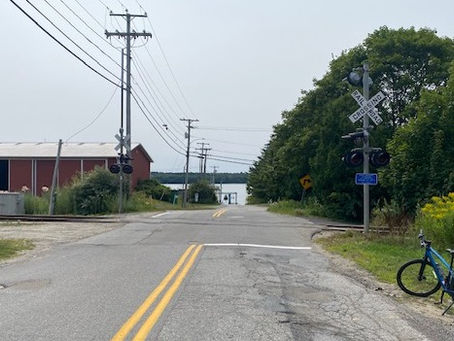 Rural road at a railroad crossing with a blue bicycle parked on the right, red building left, water visible ahead.