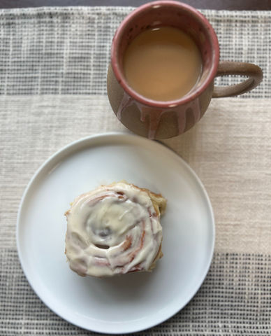 a cinnamon roll sits on a white plate next to a cup of coffee