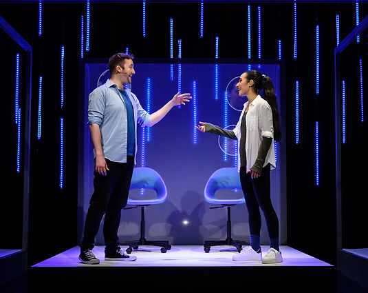 Two people look encouragingly at each other in front of a projected background on stage.