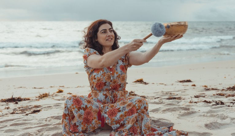 Woman in floral dress sits on a beach holding a singing bowl and mallet. Ocean waves and cloudy sky in the background, creating a serene mood.