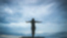 Silhouette of a person with arms outstretched against a cloudy, blue-gray sky, creating a serene and open mood atop a mountain.