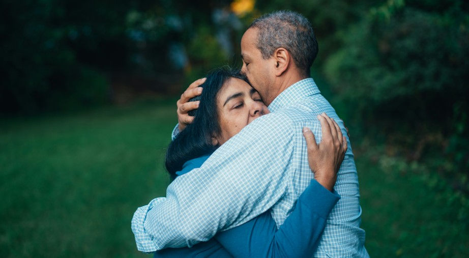 A couple hugs outdoors in a green garden, the man kissing the woman’s forehead in a tender, affectionate moment.