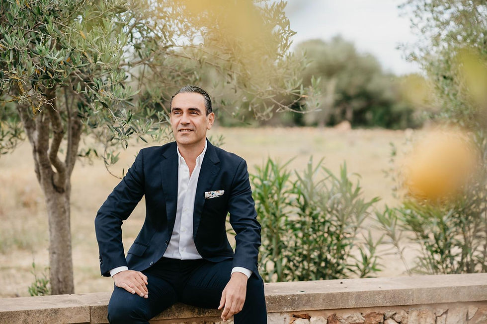 Man in a suit sits on a stone bench outdoors, surrounded by trees and greenery. He appears relaxed, with a slight smile.
