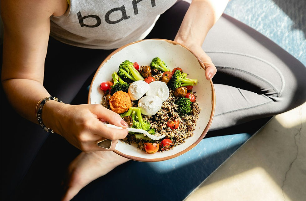 Person in a barre shirt sits on a mat, holding a bowl of quinoa, broccoli, tomatoes and mozzarella, eating with a fork.
