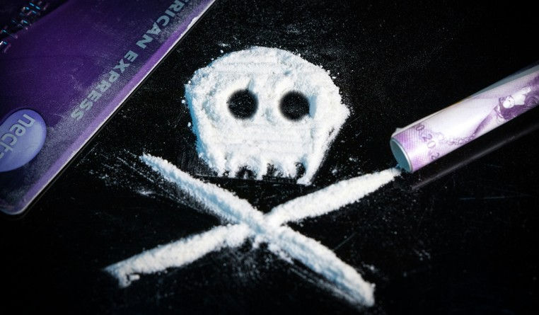 White powder forms a skull and crossbones on a black surface. A credit card and rolled note are nearby. "American Express" is visible.