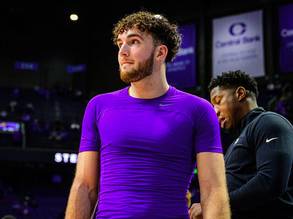 A man in a purple shirt stands in a dimly lit sports arena, with another person behind. The background includes signs and a crowd.