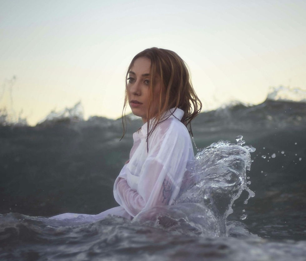 Woman in a white shirt, waist-deep in ocean waves, gazes pensively. Gray-blue water contrasts with the soft sky at dusk.