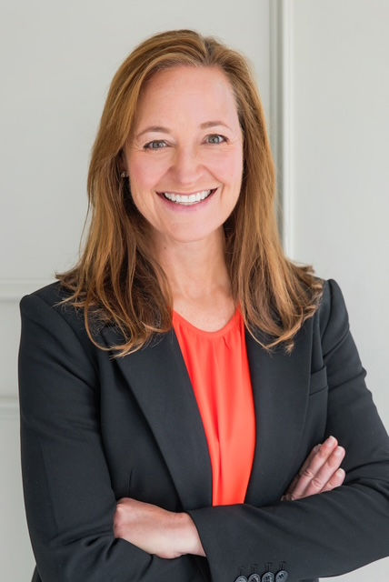 Smiling woman with brown hair in a black blazer and red top, arms crossed, standing against a light grey background.