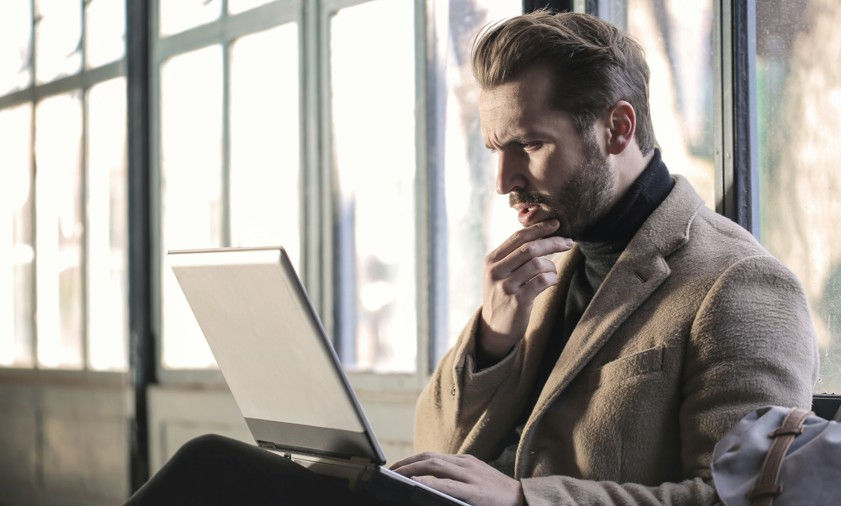Man in a beige coat sits indoors, thoughtfully looking at a laptop. Large windows create a bright, contemplative atmosphere.