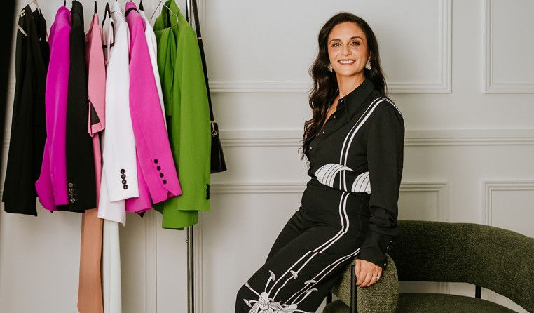Woman smiling, sitting on a green chair, in a room with a rack of colorful blazers. She's wearing a black outfit with white patterns.