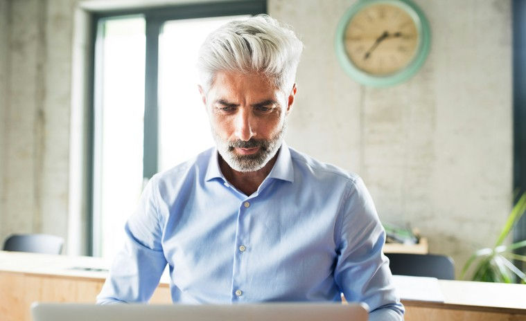 Man with gray hair in light blue shirt working on a laptop in an office. Neutral expression. Wall clock in background, natural light.