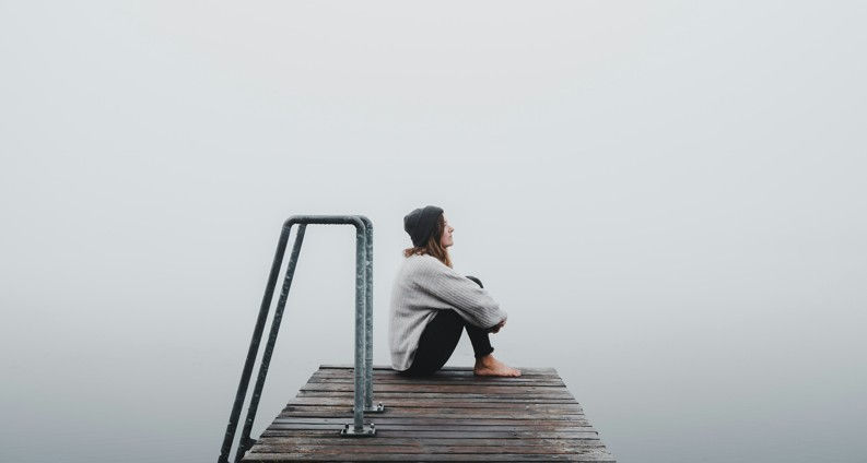 A person sits on a foggy dock, wearing a gray sweater and beanie. They look contemplative, surrounded by mist and calm water.