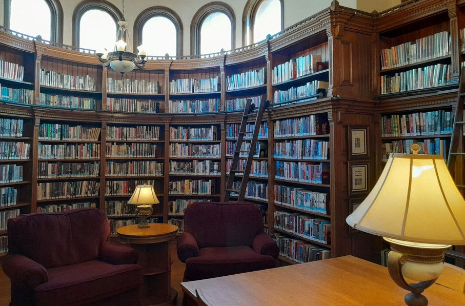 Cozy circular library with tall bookshelves, burgundy armchairs, lit lamps, and a ladder under arched windows.