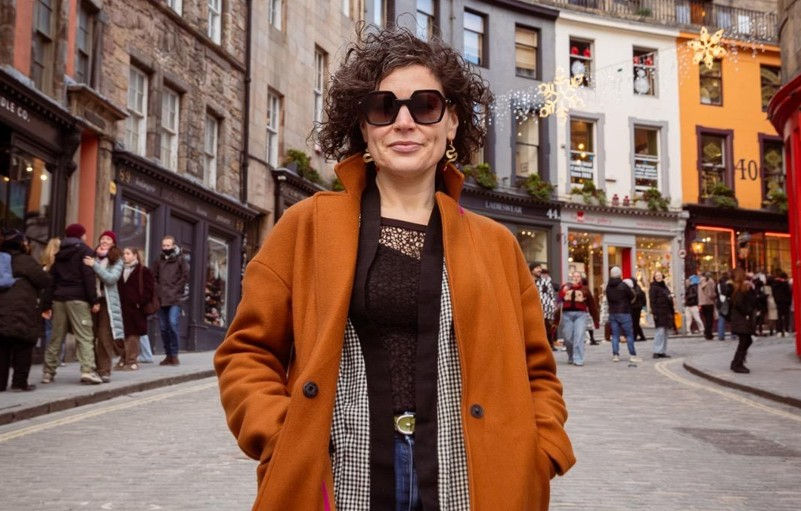 Woman in an orange coat and sunglasses stands confidently on a busy, festive street with decorated buildings and cobblestone paths.