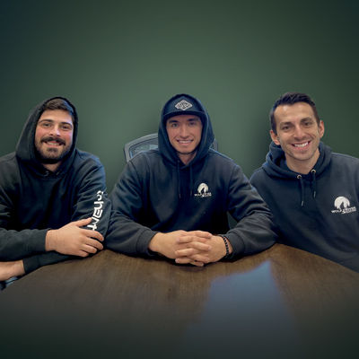 Three people in black hoodies sit at a table, smiling warmly. The background is a dark green, creating a friendly, relaxed atmosphere.