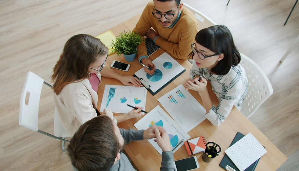 Four coworkers discuss colorful charts at a wooden table in a bright office, with phones, notebooks, and a small plant.