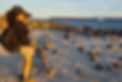 Woman in a cap and shorts photographing the ocean at sunset on a rocky beach, with distant islands and boats on the horizon.