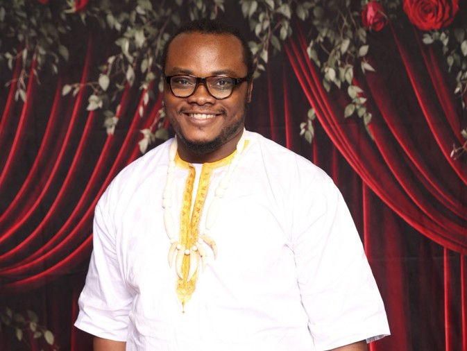 Man in white and yellow attire smiles against a backdrop of red drapes and foliage. Warm and inviting mood.