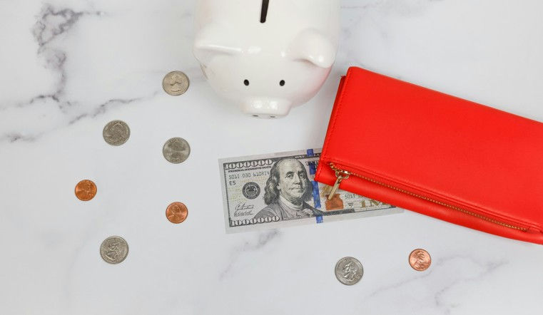 Piggy bank, coins, and a $100 bill on a marble surface. Red wallet nearby. The scene suggests saving and financial management.