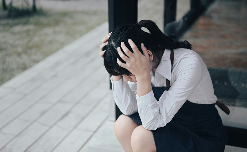 A person in a white shirt and black dress sits on a porch, holding their head in distress. The background is a blurred grassy area.
