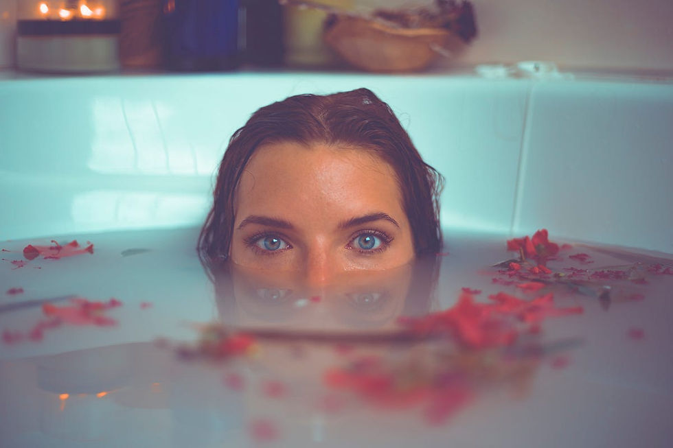 A woman's eyes peer above milky bathwater filled with red petals. Soft candlelight in the background creates a serene mood.