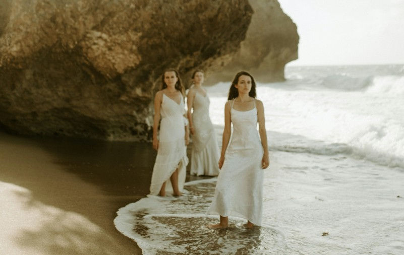 Three women in white dresses walk along a sandy beach with ocean waves. A large rock forms the background, creating a serene atmosphere.