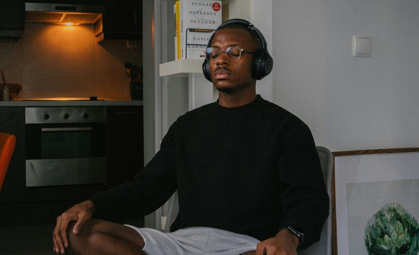 Man meditating cross-legged indoors with headphones and glasses, in a black sweater, with a warm-lit kitchen and books behind him.