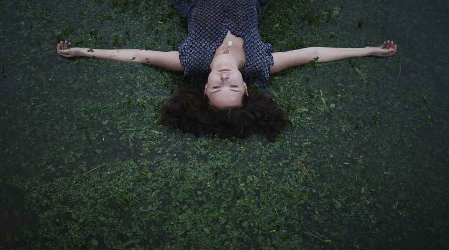 Woman floats on dark green water with arms outstretched, eyes closed, surrounded by floating leaves.