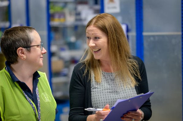 Two women are talking and smiling. One wears a green shirt; the other holds a purple clipboard. Background shows blue shelves in an office.