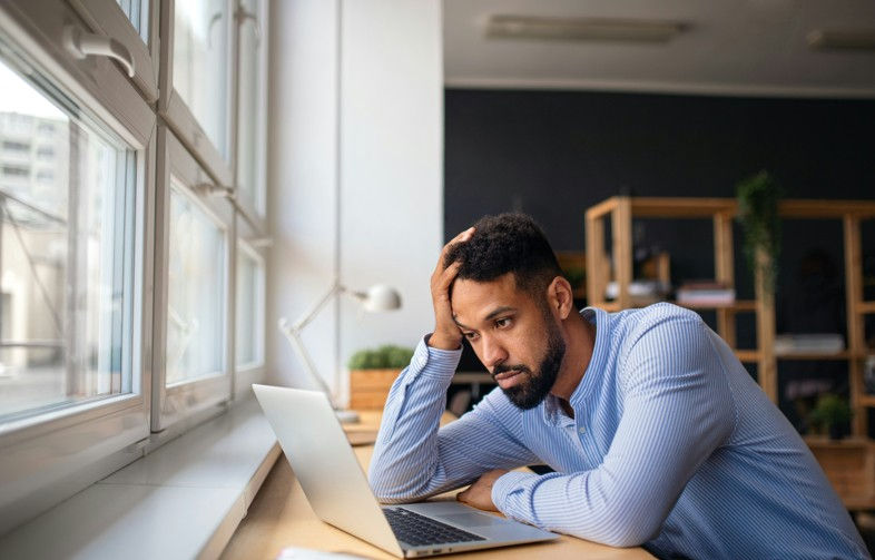 Man in blue shirt looks stressed while working on a laptop by a window. Bright office setting with plants and shelves in the background.