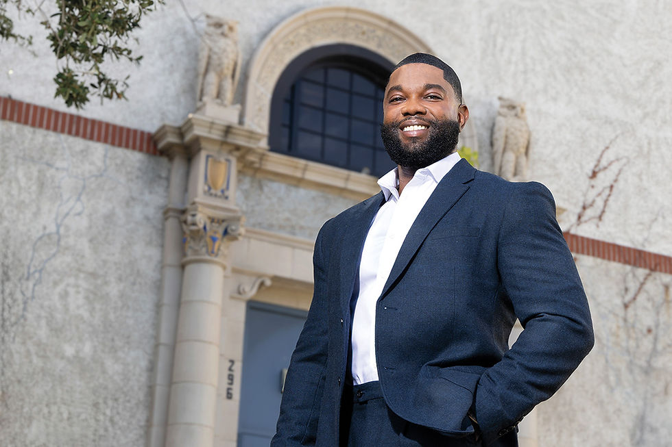 Smiling man in navy suit and white shirt stands outside an ornate stone building with arched windows.