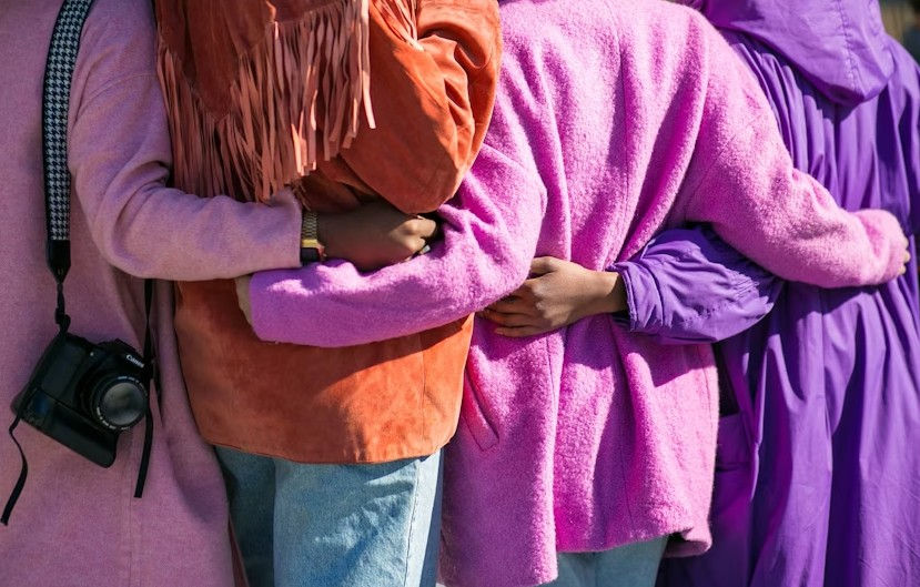 Four people in colorful coats, arms around each other. Pink, orange, and purple dominate. A camera hangs from one person's shoulder. Cozy mood.