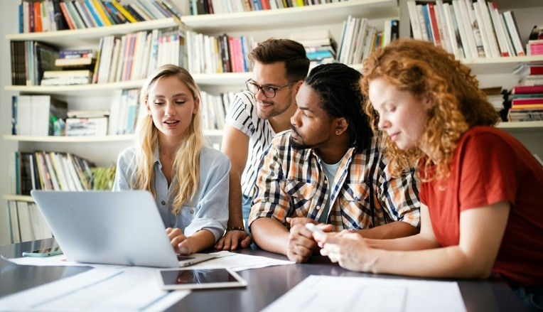 Four people collaborate around a laptop in a library, surrounded by bookshelves. The mood is focused and engaged.