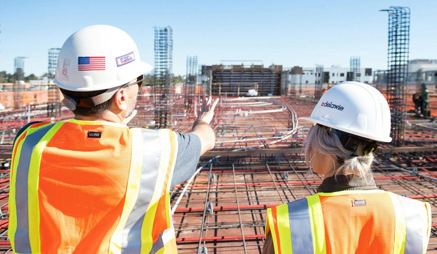 Two construction workers in safety vests and helmets discuss a building site. Rebar and building frameworks visible. Clear blue sky above.
