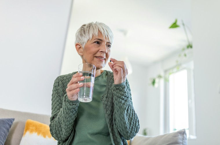 Elderly woman with short white hair holding a glass of water, about to take a pill. She sits on a sofa in a well-lit living room.