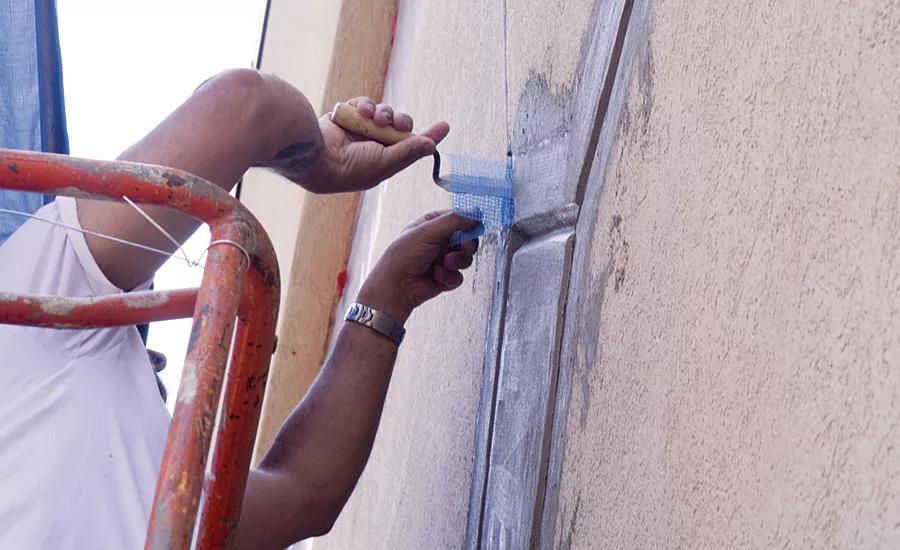 Person on scaffolding applies blue mesh to a stucco wall corner. The scene is sunny, with a focus on repair work and craftsmanship.