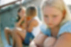 A girl in blue is sitting alone, looking sad, while two others whisper on stairs in the background. Sunlit, outdoor setting.