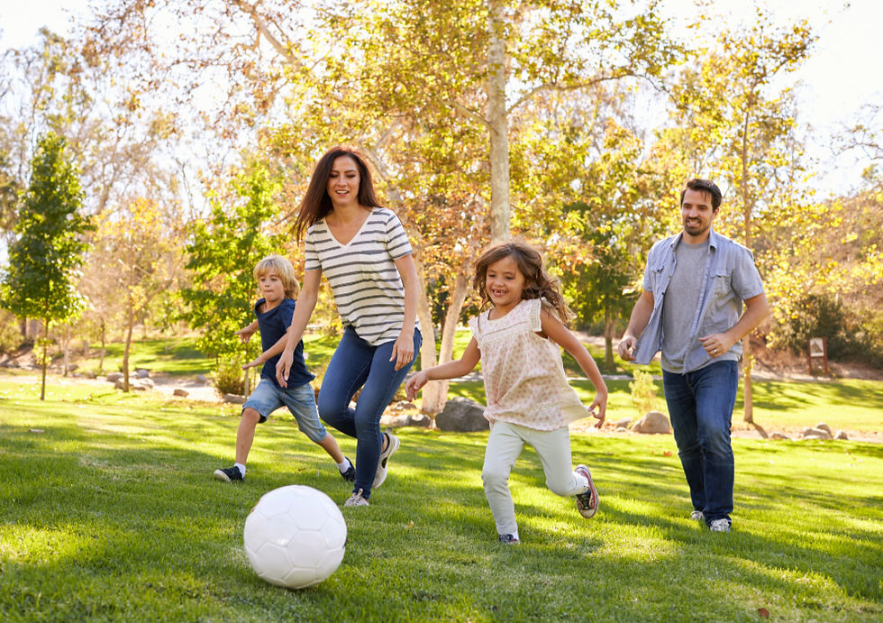 Family playing soccer in sunny park, two adults and two kids happily running on green grass with trees in the background.