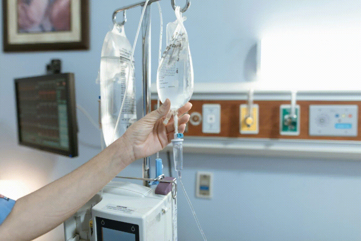 Kórházi szoba; kéz infúziós zsákot igazít; háttérben monitor; fali panelek és kapcsolók láthatók; klinikai környezet.