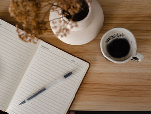 Open notebook titled "Reflection," pen resting on it, next to a vase and a cup of coffee on a wooden table | NKMT