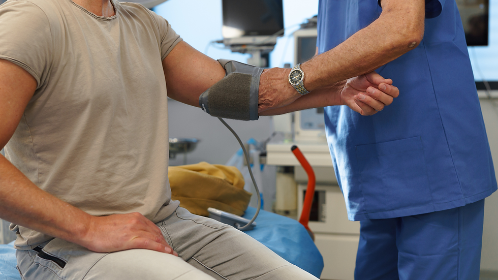 Person in beige shirt getting blood pressure taken by a healthcare worker in blue scrubs in a clinic | NKMT