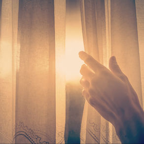Main entrouvrant un rideau léger devant une fenêtre, laissant entrer une lumière douce et dorée dans un espace intérieur calme.