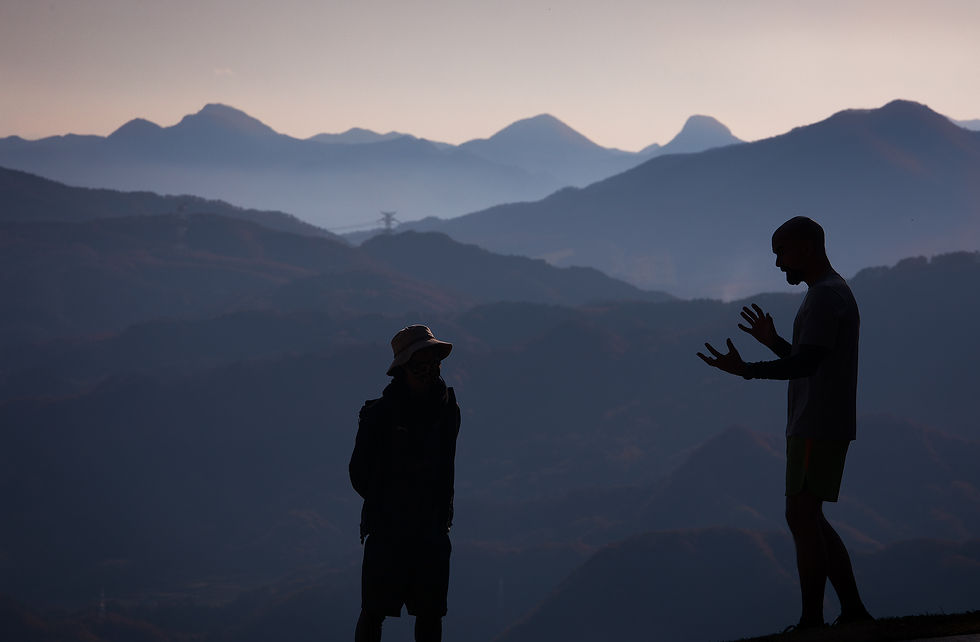 Two silhouetted figures converse with expressive gestures against a backdrop of layered mountains at dusk, evoking a serene mood.